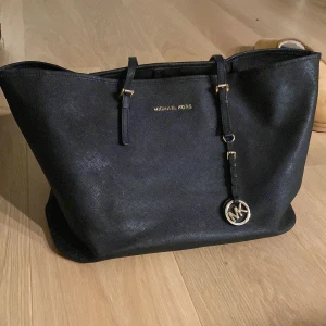 Svart axelväska från Michael Kors - Snygg svart axelväska från Michael Kors i skinn. Väskan har en stilren design med guldfärgade detaljer och en praktisk dragkedja inuti. Perfekt för att bära dina dagliga nödvändigheter med stil. Synligt använd, därav det billiga priset