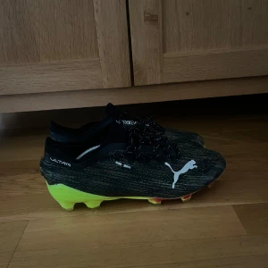 Svarta fotbollsskor från Puma - Snygga svarta fotbollsskor från Puma med vita detaljer och en färgglad sula i gult och rött. Skorna har snörning och är designade för optimal prestanda på planen. Utan snören!!!