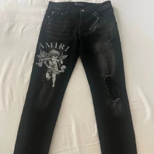 Svarta jeans med tryck från Amiri - Snygga och såklart äkta svarta jeans här från Amiri med ett unikt ängeltryck på framsidan. Jeansen har en slim passform och är dekorerade med slitningar för en trendig look. Perfekt för dig som gillar dyra märken. 