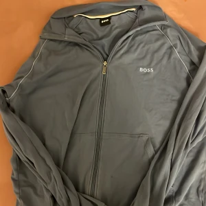 Grå jacka från Boss - Snygg grå jacka från Boss med dragkedja framtill. Jackan har en stilren design med diskret logga på bröstet och kontrastsömmar. Perfekt för en avslappnad look.