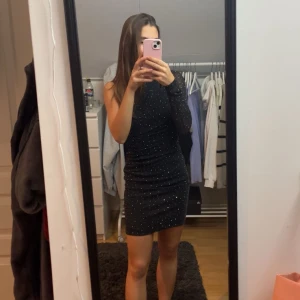 Svart glittrig klänning - Snygg svart klänning med glittriga detaljer. Den är tight och kort, perfekt för en utekväll. Klänningen en lång ärm och en figurnära passform.