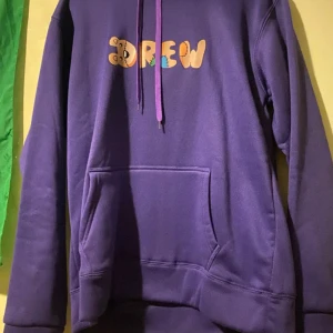 Lila hoodie från Drew - Säljer en snygg lila hoodie från Drew med en stor orange smiley på ryggen och texten 'Drew' på framsidan. Hoodien har en klassisk passform med långa ärmar och en praktisk ficka framtill. Perfekt för en avslappnad stil!