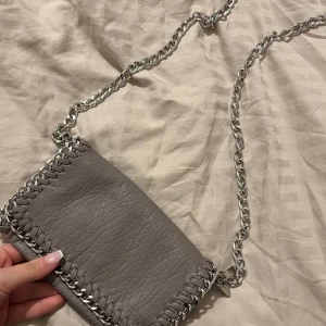 Grå axelväska med kedjedetaljer - Snygg grå axelväska från Tiamo med silverfärgad kedja och dekorativa sömmar. Perfekt för att ge din outfit en stilfull touch. Väskan har en praktisk dragkedja för säker förvaring. Den är knappast använd, pris kan diskuteras