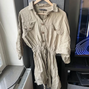 Beige klänning second hand - Säljer en stilren linneklänning med knappar framtill och två bröstfickor. Klänningen har en avslappnad passform med resår i midjan och långa ärmar. Perfekt för en casual look!
