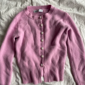 Rosa kofta från Zara - Säljer en söt rosa kofta från Zara med dekorativa pärlknappar. Koftan är långärmad och har en ribbad kant vid halsen och ärmsluten. Perfekt för att lägga till en färgklick i garderoben!