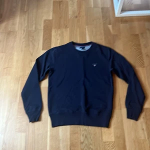 Mörkblå tröja från GANT - Säljer en stilren mörkblå tröja från GANT med broderad logotyp på bröstet. Tröjan har långa ärmar och ribbade muddar vid ärmslut och nederkant. Perfekt för en avslappnad och snygg look.