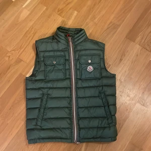 Grön dunväst från Moncler - Snygg grön dunväst från Moncler med dragkedja och två fickor med knappar framtill. Perfekt för lager-på-lager under kyliga dagar. Moncler-logga på bröstet ger en exklusiv touch.
