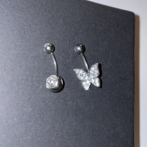 Silver navelpiercing med fjäril och kristall - Snygga navelpiercingar i silver med en glittrande fjäril och en klar kristall. Perfekt för att ge lite extra bling till din stil. Fjärilen är täckt med små gnistrande stenar som fångar ljuset vackert. De är rengjorda men kokat vatten. 70 kr styck, båda för 115 kr.