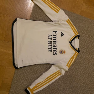 Vit långärmad Real Madrid tröja från Adidas - Säljer en vit långärmad Real Madrid tröja från Adidas med gula ränder på ärmarna och svart krage. Tröjan har klubbens emblem och sponsorlogga på framsidan. Perfekt för fotbollsfans!