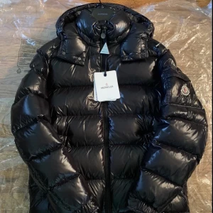 Svart dunjacka från Moncler - Snygg och glansig svart dunjacka från Moncler med huva och dragkedja. Perfekt för kalla dagar med sin pufferdesign som håller värmen. Jackan har en klassisk look med Moncler-logga på ärmen.