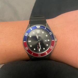 Casio klocka - Snygg Casio klocka med svart urtavla och datumvisning. Bezel i blått och rött ger en sportig look. Armbandet är i svart material och har ett klassiskt spänne. Perfekt för både vardag och äventyr.