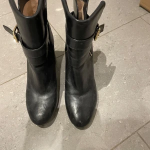 Svarta boots med gulddetaljer från Michael Kors - Snygga svarta boots i skinn med guldfärgade spännen. De har en klack och en stilren design som passar perfekt för höst och vinter. Perfekta för att ge din outfit en elegant touch.