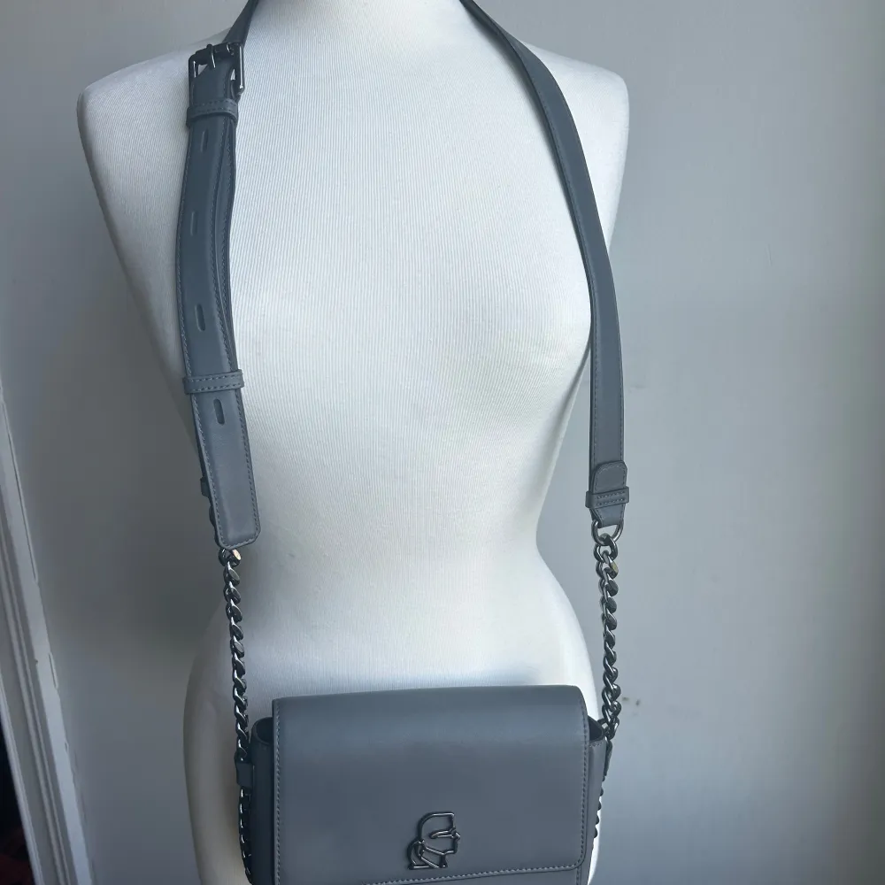 Säljer en Karl Lagerfeld stilren grå axelväska med en cool kedjedetalj på axelremmen. Väskan har en elegant design med ett metallmärke på framsidan. Perfekt för både vardag och fest!. Laukut & Käsilaukut.