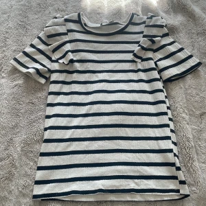 Randig topp med volangdetaljer - Säljer en superfin randig topp i svart och vit med volangdetaljer på axlarna. Den har korta ärmar och är perfekt för alla tillfällen. Materialet känns mjukt och bekvämt.