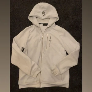 Sailracing bowman zip hoodie  - Vit zip hoodie från sailracing. Väl använd och har därför några få fläckar som syns i viss ljus. Orginalpris 1.500kr. Storlek 160 och passar på både tjej och kille. säljer pga av att jag har växt ut den. Skriv för fler bilder!