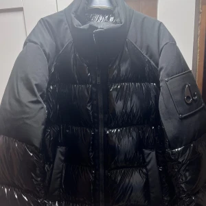 Svart pufferjacka från Moose Knuckles - Säljer en svart pufferjacka från Moose Knuckles. Jackan har en glansig finish och är perfekt för vintern. Den har en dragkedja framtill och en cool detalj på ärmen. Insidan är quiltad för extra värme. Perfekt för vintern! Pris kan diskuteras!!