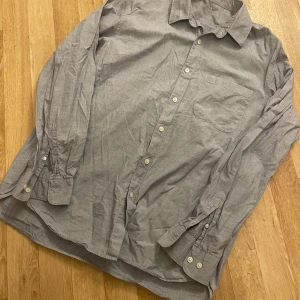 Blå långärmad skjorta - Säljer en stilren grå skjorta med långa ärmar och klassisk krage. Skjortan har knappar framtill och en bröstficka. Perfekt för en avslappnad men ändå uppklädd look.