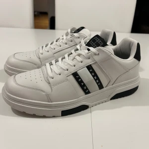 Vita sneakers från Tommy Jeans (använd 1 gång)  - Snygga vita sneakers från Tommy Jeans med svarta detaljer och snörning. Skorna har en stilren design med perforeringar på tån för extra ventilation. Perfekta för en casual look. 