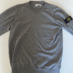 Grå tröja från Stone Island Junior - Säljer en stilren grå tröja från Stone Island Junior. Tröjan har en rund halsringning och ett ikoniskt märke på ärmen. Perfekt för en avslappnad och trendig look.