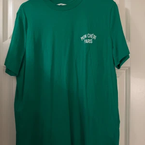 Grön t-shirt med tryck 'Mon Chéri Paris' - Snygg grön t-shirt med trycket 'Mon Chéri Paris' på bröstet. Perfekt för en avslappnad stil. T-shirten har en klassisk rund hals och korta ärmar.