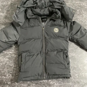 Svart pufferjacka från Trapstar - Säljer en mörkgrå pufferjacka från Trapstar med huva och dragkedja. Jackan har ett stort Trapstar-tryck på ryggen och en rund logotyp på bröstet. Perfekt för kalla dagar med sin vadderade design. Bara använd några gånger och säljer den eftersom den är för stor för mig. 