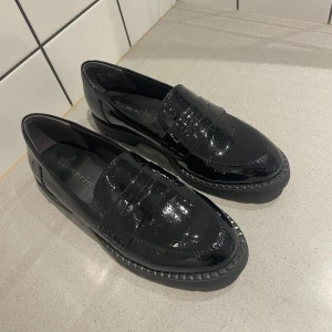 Svarta lackloafers från Tamaris - Snygga svarta loafers i lack från Tamaris använda ENDAST 1 gång. Skorna har en klassisk design med en låg klack och en robust sula. Perfekta för en stilren look. Säljer då jag fick dem i present och de är tyvärr fel storlek för mig. Storlek 38. Nypris 999 kr!