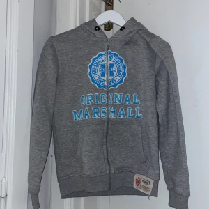 Grå hoodie - Grå hoodie från Marshall Original med dragkedja och blå text och märke på framsidan. Har även blåa snören som inte är i men som man kan få med🩷