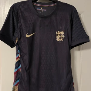 Englands fotbollströja - Snygg svart fotbollströja från Nike med korta ärmar och ett stilrent märke i guld på bröstet. Tröjan har en diskret randig textur och färgglada detaljer på sidorna. Perfekt för fotbollsfans som vill ha något unikt och trendigt.