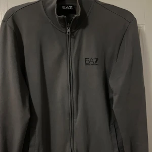 Grå tröja från EA7 Armani  - Snygg grå jacka från EA7 Armani med dragkedja framtill och diskret logga på bröstet. Perfekt för en stilren look. Jackan har långa ärmar och en enkel design som passar till det mesta.