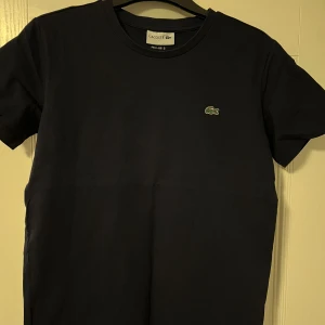 Svart t-shirt från Lacoste - Säljer en stilren svart t-shirt från Lacoste med den klassiska krokodilloggan på bröstet. T-shirten är kortärmad och gjord i mjuk bomull. Perfekt för en avslappnad look.