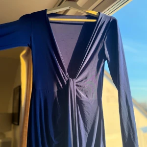 Mörkblå långklänning med v-ringning från NAKD - Elegant mörkblå långklänning med djup v-ringning och långa ärmar. Perfekt för en stilfull kväll. Klänningen har en smickrande passform och är tillverkad i ett mjukt material som ger ett fint fall. NAKD