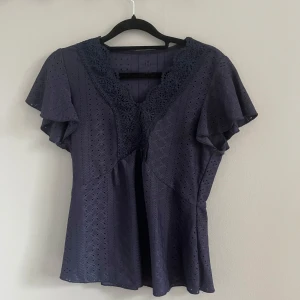 Mörkblå blus med spetsdetaljer - Säljer en elegant mörkblå blus med vackra spetsdetaljer vid halsringningen. Blusen har korta ärmar med volang och ett diskret hålmönster över hela tyget. Perfekt för en stilren look. 