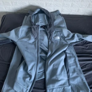 Grå jacka från The North Face - Säljer en grå tracksuit från The North Face med dragkedja och huva. Jackan har en stilren design med logotyp på bröstet. Perfekt för kyligare dagar och passar bra till både träning och vardag. Byxorna följer även med så att man kan gå runt med hela dressen. Koftan är använd ca 4 gånger och byxorna är bara testade en gång. Inga priser är spikade går självklart att förhandla. Hela dressen är köpt för ca 2500 på jd sport