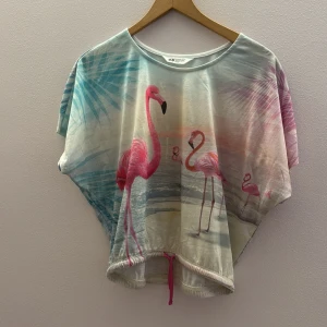 Färgglad topp med flamingomotiv från H&M - Säljer en färgglad topp från H&M med ett tropiskt flamingomotiv. Toppen har korta ärmar och en lös passform med ett knytband i nederkanten. Perfekt för en somrig look!