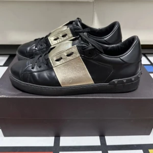 Svarta sneakers med gulddetaljer - Snygga svarta sneakers från Valentino med guldfärgade detaljer på sidorna. Skorna har snörning och en stilren design med en mönstrad sula för extra grepp. Perfekta för en trendig look.