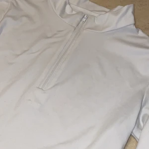 Vit långärmad tröja med dragkedja - Vit långärmad tröja med dragkedja framtill. Perfekt för en sportig look eller som ett basplagg i garderoben. Tröjan har en croppad passform och är tillverkad i ett mjukt material.