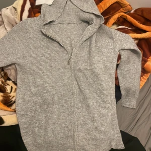 Grå ribbad hoodie - Säljer en grå kashmir hoodie med dragkedja. Den har långa ärmar och en bekväm huva, perfekt för kyliga dagar. Passar bra till både avslappnade och aktiva tillfällen. (Pris kan diskuteras) helt nytt skick förekommer i paket. Ej använd då den är stor på mig
