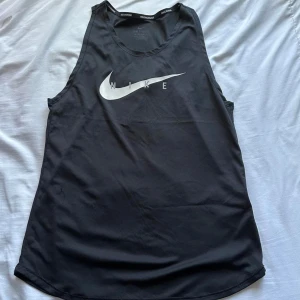 Svart Nike swoosh linne - Säljer en svart linnetopp från Nike med det klassiska logotyptrycket i vitt på framsidan. Toppen är ärmlös och har en sportig design, perfekt för träning eller en aktiv livsstil. 