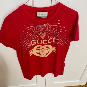 Röd t-shirt från Gucci med strassdetaljer - Snygg röd t-shirt från Gucci med glittrande strassdetaljer och det ikoniska Gucci-logotypen i guld på framsidan. Använd en gång och tvättad en gång. Storlek M 