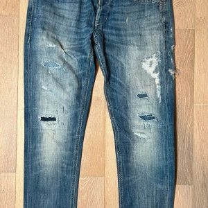 Dondup jeans - Säljer ett par blå dondup jeans med slitningar och hål för en trendig look. Jeansen har en normal passform och är tillverkade i denim. Perfekta för en avslappnad stil.
