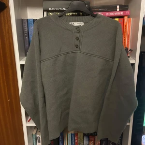 Grå tröja från Zara - Säljer en stilren grå tröja från Zara med knappar vid halsen. Tröjan har långa ärmar och en ribbad kant vid ärmslut och nederkant. Perfekt för en avslappnad look.