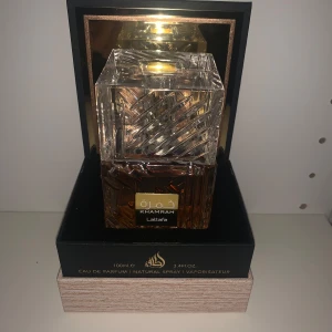 Khamrah Eau de Parfum från Lattafa - Elegant parfymflaska med en lyxig design i glas. Flaskan har en fyrkantig form med ett mönstrat lock och en gyllene etikett. Parfymen har en varm bärnstensfärg och kommer i en stilren svart och guld kartong. Perfekt för den som söker en sofistikerad doftupplevelse.