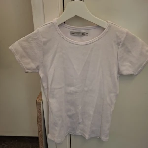 Vit ribbad t-shirt - Säljer en enkel och stilren vit ribbad t-shirt från Lager 157. Den har en rund halsringning och korta ärmar, perfekt för en avslappnad look. Passar bra till jeans eller kjol.