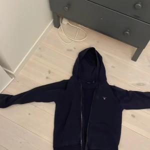 Mörkblå hoodie från Gant  - Säljer en stilren mörkblå hoodie från Gant med dragkedja framtill. Perfekt för kyliga dagar med sin mjuka och bekväma passform. Den har en klassisk design med huva och långa ärmar den är storlek 158-164 men den är väldigt liten för sin storlek 