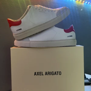 Vita sneakers från Axel Arigato - Snygga vita sneakers från Axel Arigato med röd detalj vid hälen och texten '1.40MM' på sidan. Skorna har en klassisk design med snörning och en stilren vit sula. Perfekta för en stilren look.