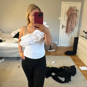 Vit Zara topp med volang - Säljer en stilren vit oneshoulder topp med volangdetalj. Perfekt för en trendig look. Toppen har en åtsittande passform och är tillverkad i ett mjukt material. Använd fåtal gånger💞💞💞
