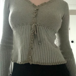 Beige ribbad kofta med knyt - Säljer en beige/grönish ribbad kofta med knyt och knappar framtill. Koftan har långa ärmar och en v-ringad design. Den är lite glittrig! Hör av dig om frågor uppstår ❤️