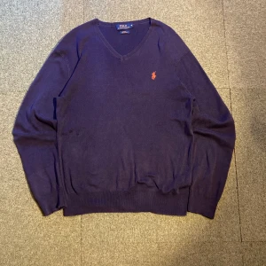 Mörkblå tröja från Ralph Lauren - Säljer en stilren mörkblå tröja från Ralph Lauren i Pima bomull. Tröjan har en v-ringad design och ett broderat orange logotyp på bröstet. Perfekt för en elegant och avslappnad look. Passar som medium vilket det också står på taggen, funkar perfekt på mig och jag är 180 cm och väger ca 60 kg