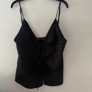 Svart satinlinne med knytband - Snygg svart satinlinne från Zara med smala axelband och knytband framtill. Perfekt för en stilren look. Passar bra till både jeans och kjol.