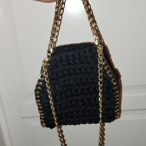 Svart virkad handväska med guldkedja - Snygg svart virkad handväska med en elegant guldkedja. Perfekt för att ge din outfit en trendig touch. Väskan har en unik textur och är både stilren och praktisk.